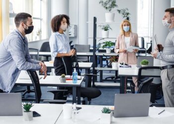 Modern team meeting, group work and social distancing. Manager with tablet speaks with workers in protective masks in interior of modern office with gadgets during coronavirus epidemic, free space