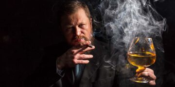 Solid confident bearded man in suit with glass of whisky and cigar with fume studio portrait