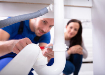 Young Woman Looking At Male Plumber Fixing Sink Pipe In Kitchen