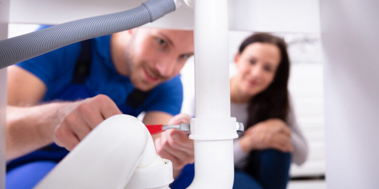 Young Woman Looking At Male Plumber Fixing Sink Pipe In Kitchen