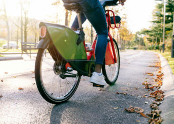 rear shot photograph of a student boy with a bag on his back carrying a red shared electric bike along a tree-lined park path at sunset