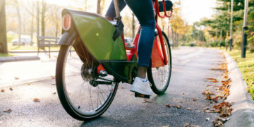 rear shot photograph of a student boy with a bag on his back carrying a red shared electric bike along a tree-lined park path at sunset