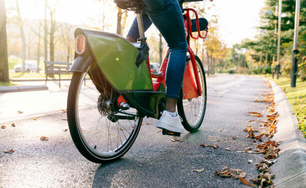 rear shot photograph of a student boy with a bag on his back carrying a red shared electric bike along a tree-lined park path at sunset