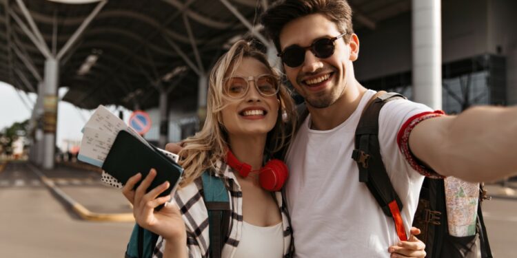 Pretty blonde woman in sunglasses, plaid shirt holds plane tickets and passport. Brunette man hugs girlfriend and takes selfie near airport. Portrait of travelers.