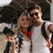 Pretty blonde woman in sunglasses, plaid shirt holds plane tickets and passport. Brunette man hugs girlfriend and takes selfie near airport. Portrait of travelers.